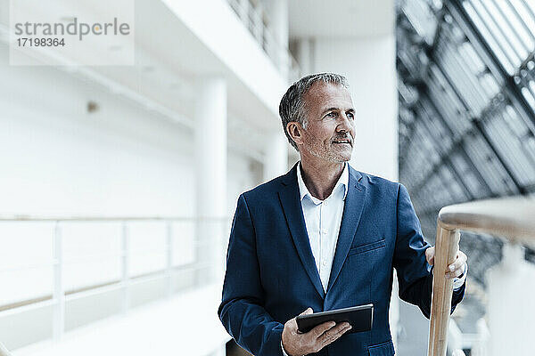 Lächelnder Geschäftsmann mit digitalem Tablet  der im Bürokorridor wegschaut