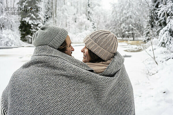 Lächelndes Paar in Decke gehüllt sitzt am gefrorenen Fluss im Wald