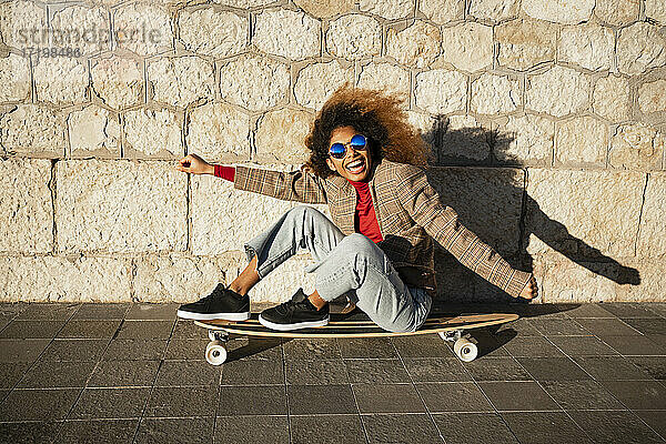 Fröhliche Frau mit ausgestreckten Armen  die auf einem Skateboard an der Wand sitzt  an einem sonnigen Tag