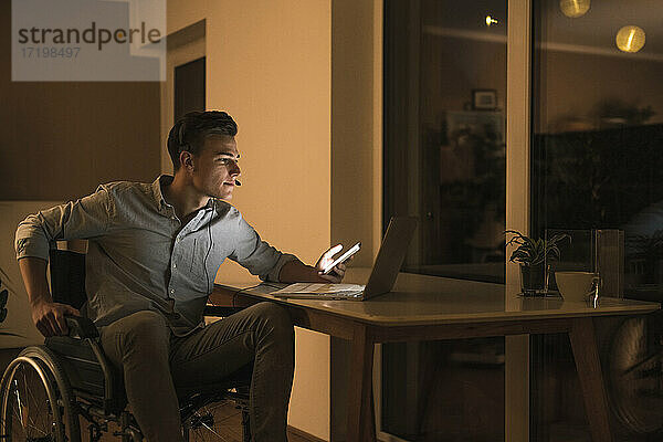 Behinderter männlicher Geschäftsmann mit Laptop und Kopfhörern  während er im Wohnzimmer im Rollstuhl sitzt