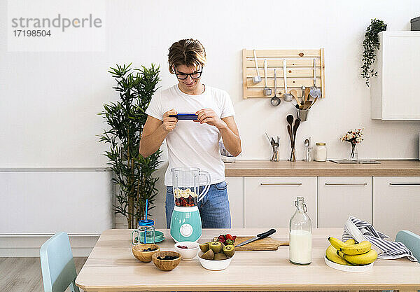 Junger Mann fotografiert Früchte im Mixer mit seinem Smartphone in der Küche zu Hause