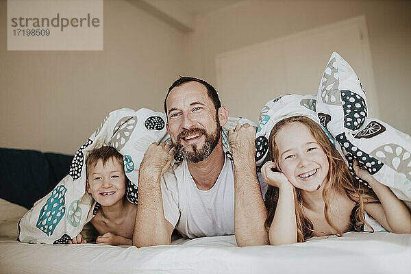 Lächelnder Vater mit Tochter und Sohn auf dem Bett liegend unter einer Decke im Schlafzimmer zu Hause
