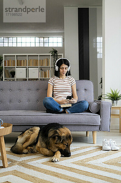 Frau  die auf dem Sofa sitzend Musik hört  während der Hund auf dem Teppich im Wohnzimmer schläft