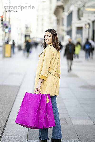 Junge Frau mit Einkaufstüten  die über die Schulter schaut  während sie auf einem Fußweg in der Stadt spazieren geht