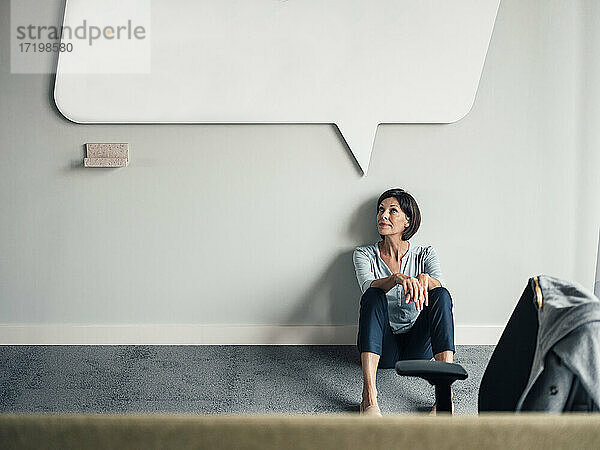 Nachdenkliche Geschäftsfrau schaut auf  während sie im Büro auf dem Boden sitzt