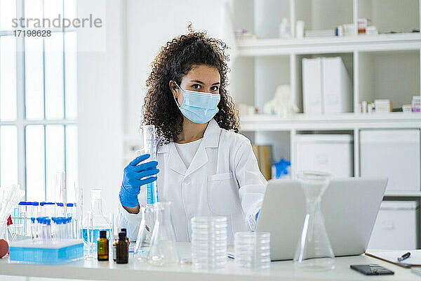 Junge Wissenschaftlerin mit Reagenzglas und Laptop im Krankenhaus während COVID-19