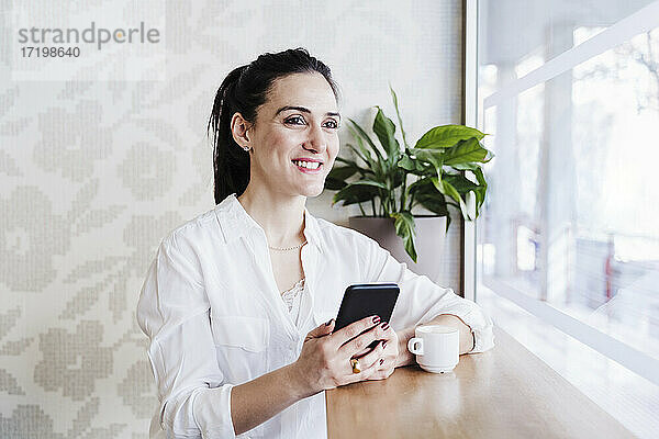 Lächelnde Geschäftsfrau mit Mobiltelefon  die durch ein Fenster im Büro schaut
