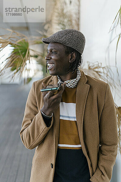 Lächelnder Mann mit flacher Kappe  der über einen Lautsprecher mit seinem Smartphone spricht