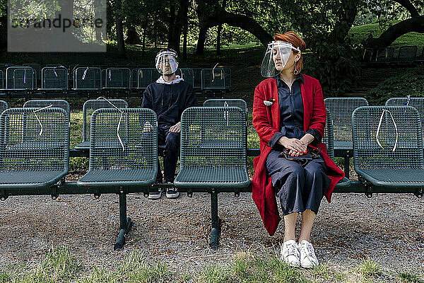 Mutter und Sohn mit Gesichtsschutz auf einem Sitz im Park während COVID-19