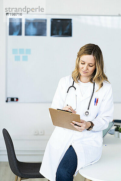 Eine Ärztin schreibt auf einem Klemmbrett  während sie auf einem Schreibtisch in einer medizinischen Klinik sitzt