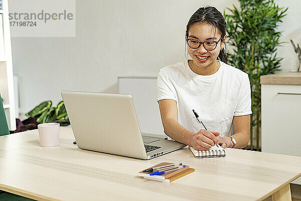 Lächelnde Frau  die in einen Notizblock schreibt  während sie zu Hause am Laptop sitzt