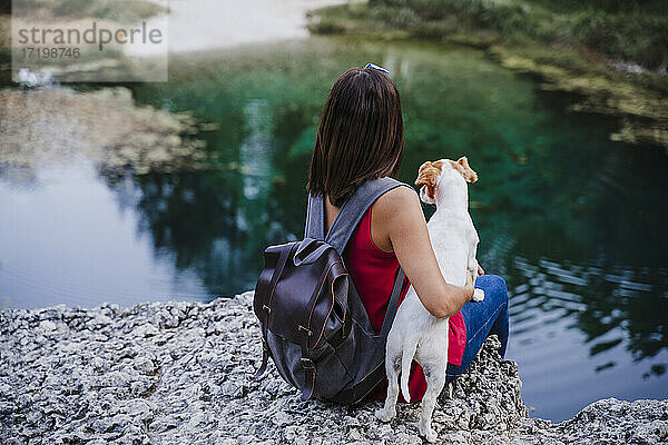 Frau mit Rucksack  die ihren Hund umarmt  während sie auf einem Felsen am See sitzt