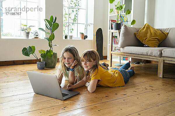 Lächelnde Freundinnen schauen auf einen Laptop  während sie im Wohnzimmer auf der Vorderseite liegen