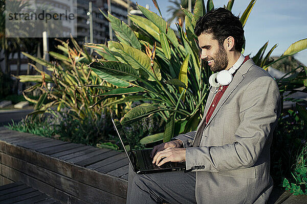 Lächelnder Geschäftsmann  der einen Laptop benutzt  während er auf einer Stützmauer sitzt