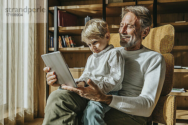 Junge  der auf dem Schoß seines lächelnden Vaters im Wohnzimmer sitzt und e-learning betreibt