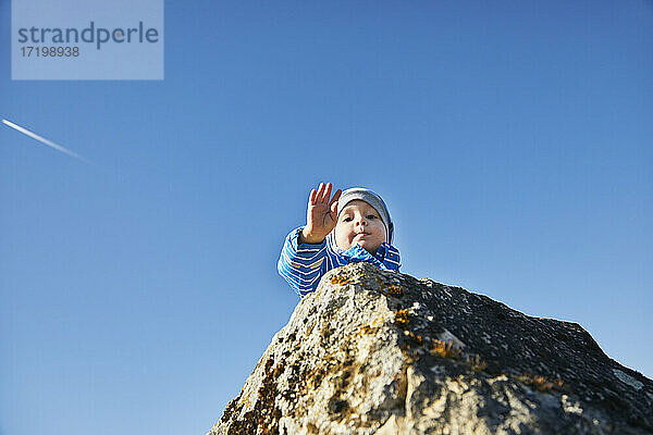 Niedlicher Junge auf einem Felsen mit winkender Hand gegen den Himmel