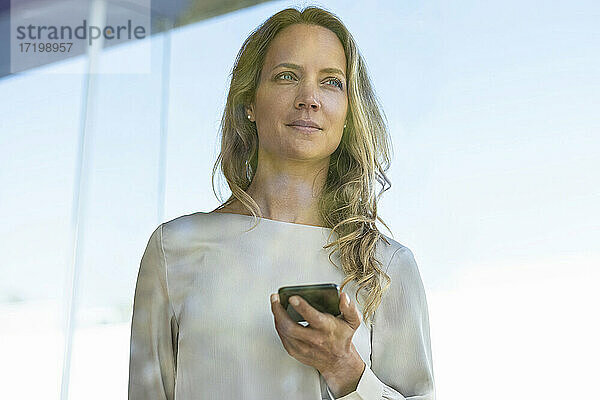Weibliche Fachkraft mit Mobiltelefon vor einem Glasfenster im Büro