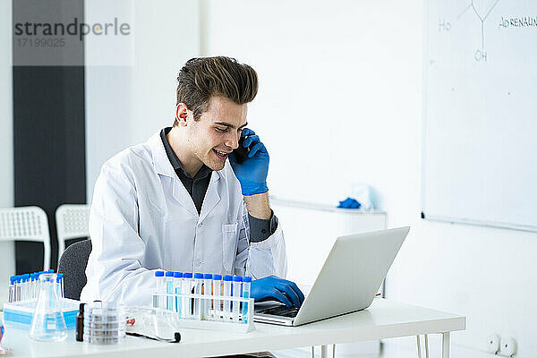Männlicher Wissenschaftler  der mit einem Smartphone spricht  während er einen Laptop am Schreibtisch im Labor benutzt