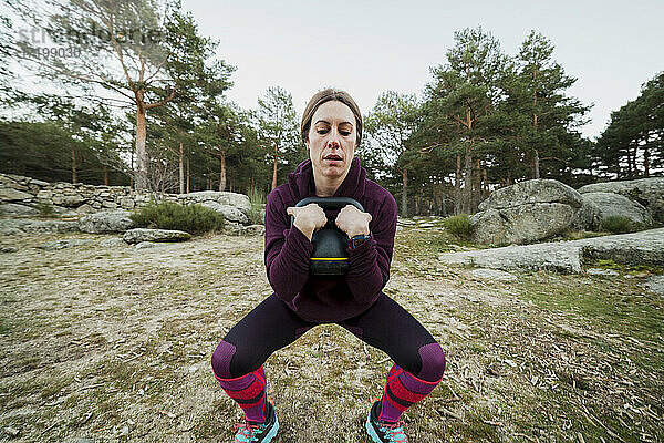 Mittlere erwachsene Frau in hockender Position bei einer Übung mit einer Kettlebell im Wald