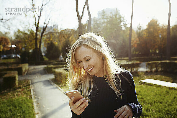 Blonde Frau lacht  während sie ihr Smartphone im Park verklagt