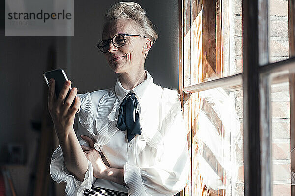 Lächelnde Geschäftsfrau  die auf ihr Mobiltelefon schaut  während sie am Fenster im Büro zu Hause steht