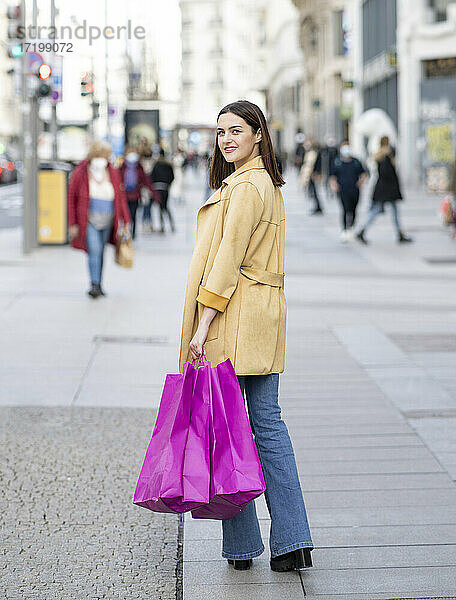Lächelnde junge Frau mit magentafarbenen Einkaufstaschen auf dem Fußweg in der Stadt