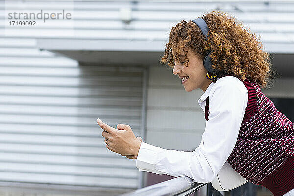 Lächelndes Teenager-Mädchen mit Kopfhörern  das ein Mobiltelefon benutzt und sich auf ein Geländer stützt