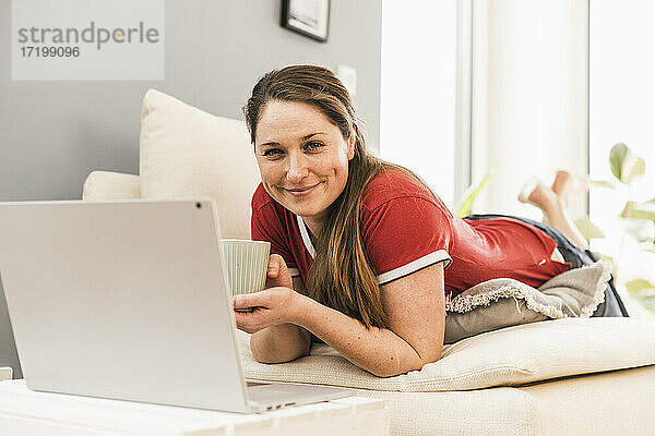 Lächelnde Frau mit Kaffeetasse auf dem Sofa zu Hause liegend