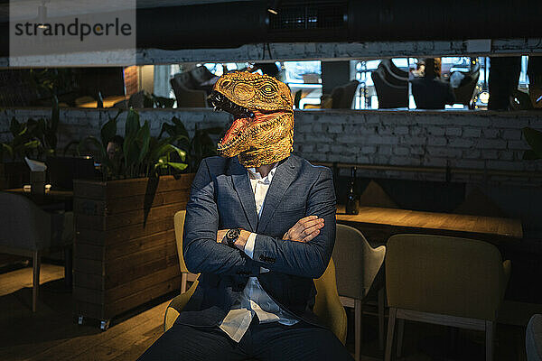 Geschäftsmann mit Dinosauriermaske  der mit verschränkten Armen in einem Café sitzt