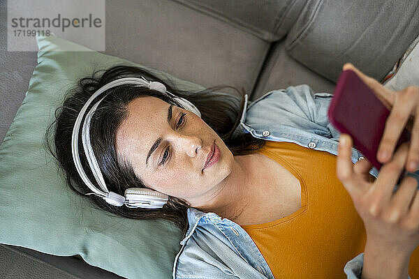 Junge Frau mit Kopfhörern  die ein Mobiltelefon benutzt  während sie zu Hause auf dem Sofa liegt