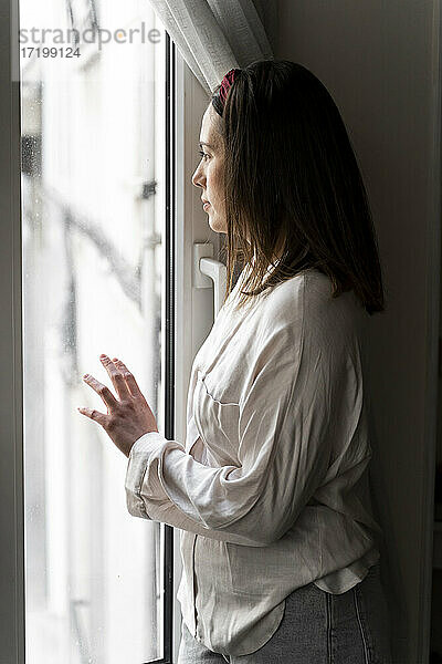 Nachdenkliche junge Frau schaut durch das Fenster  während sie zu Hause steht