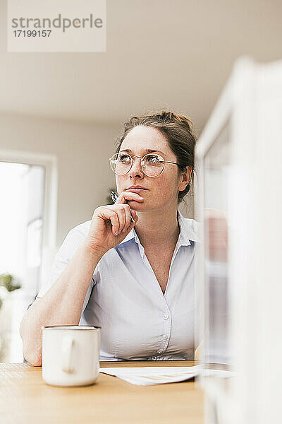 Nachdenkliche Unternehmerin mit Brille sitzt am Schreibtisch und schaut weg