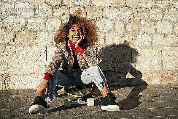 Lachende Afro-Frau  die auf einem Skateboard an der Wand sitzt  an einem sonnigen Tag