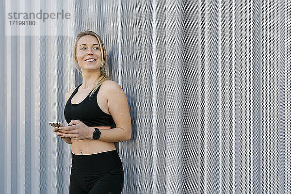 Lächelnde Sportlerin in Sportkleidung  die wegschaut und ihr Handy an die Wand hält