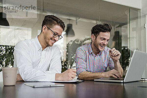 Lächelnder Geschäftsmann arbeitet am Laptop  während ein männlicher Kollege im Büro auf dem Klemmbrett schreibt