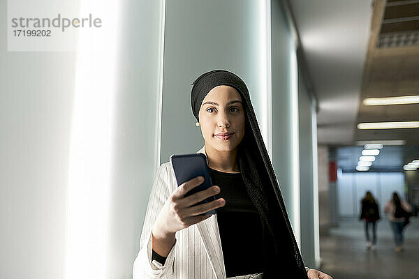 Junge arabische Frau  die ein Smartphone benutzt  während sie in der Nähe einer Wand in einer Einkaufsstraße steht
