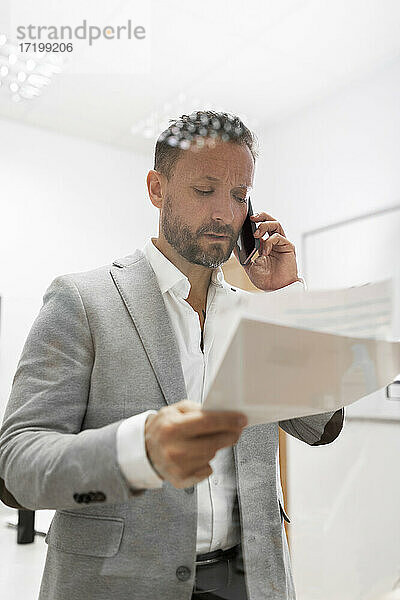 Älterer Unternehmer mit Dokumenten  der im Büro mit seinem Smartphone telefoniert