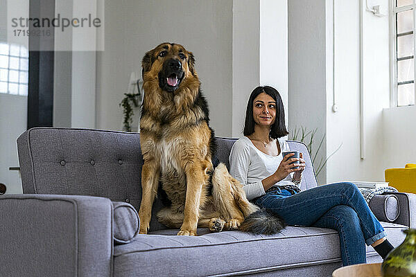 Lächelnde Frau mit Kaffeetasse  die den Hund ansieht  während sie auf dem Sofa im Wohnzimmer sitzt