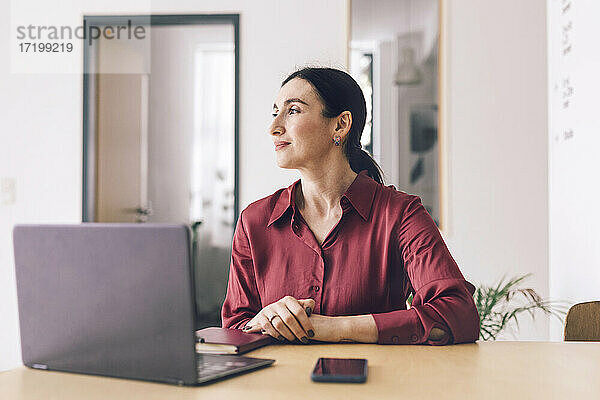 Lächelnde Geschäftsfrau mit Laptop  die wegschaut  während sie am Tisch im Heimbüro sitzt