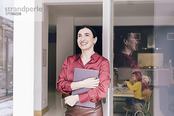 Lächelnde Geschäftsfrau  die wegschaut  während sie einen Laptop an einer Glastür im Haus hält