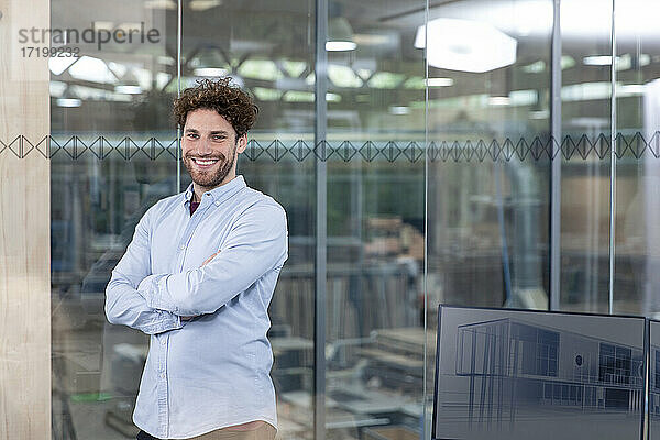 Lächelnder männlicher Unternehmer  der sich im Büro an eine Glaswand lehnt