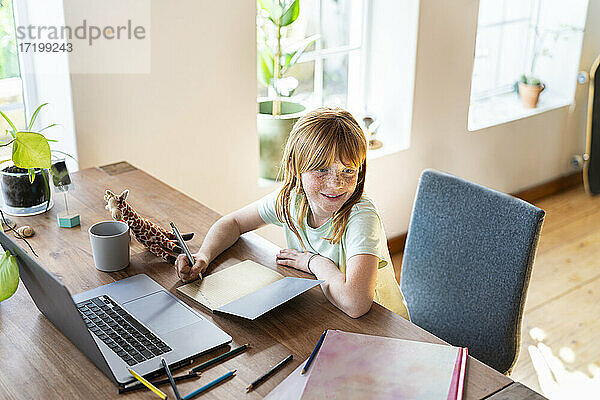 Lächelndes rothaariges Mädchen  das wegschaut  während es zu Hause vor dem Laptop Hausaufgaben macht