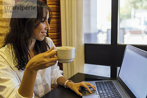 Junge Geschäftsfrau arbeitet am Laptop und hält eine Tasse Kaffee in einem Café