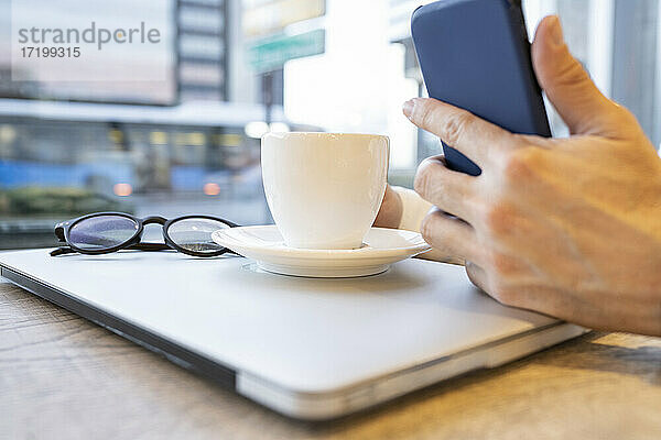 Die Hand eines Geschäftsmannes  der ein Smartphone benutzt  während er einen Kaffee mit einem Laptop am Kaffeetisch trinkt