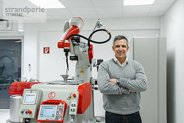 Lächelnder Geschäftsmann mit verschränkten Armen vor einer Maschine in einer Fabrik