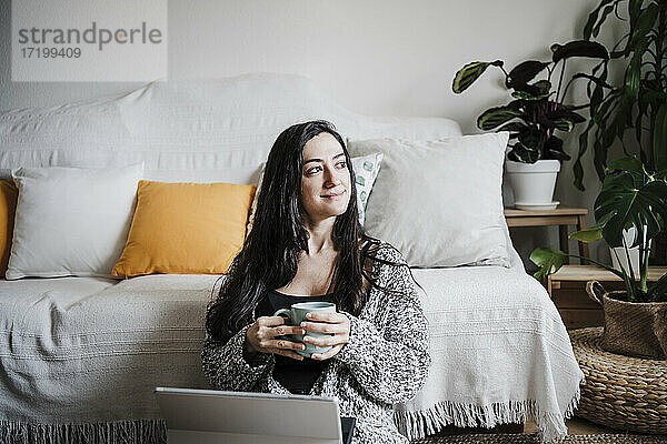 Lächelnde Frau mit Laptop  die eine Kaffeetasse hält und auf dem Boden vor dem Sofa im Wohnzimmer sitzt