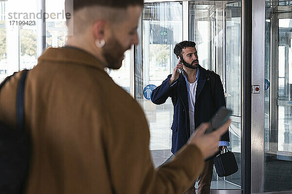 Geschäftsmann  der mit einem Mobiltelefon spricht  während er mit einem Mitarbeiter im Hintergrund vor einem Gebäude steht