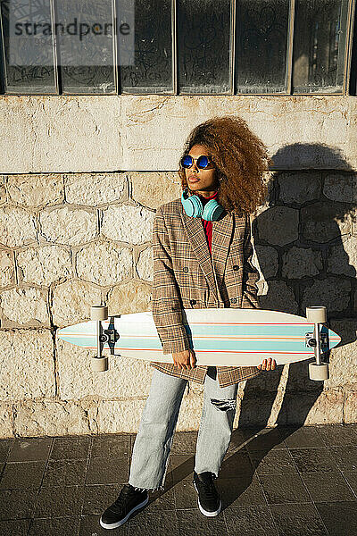 Coole Einstellung Frau trägt Sonnenbrille mit Skateboard stehen gegen Gebäude während sonnigen Tag