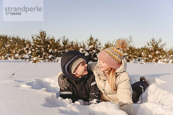 Verspielte Geschwister auf Schnee liegend gegen den Himmel im Winter