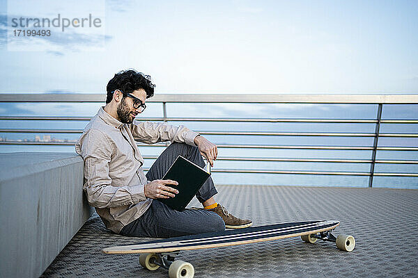 Geschäftsmann  der Tagebuch liest  während er mit seinem Longboard am Pier sitzt